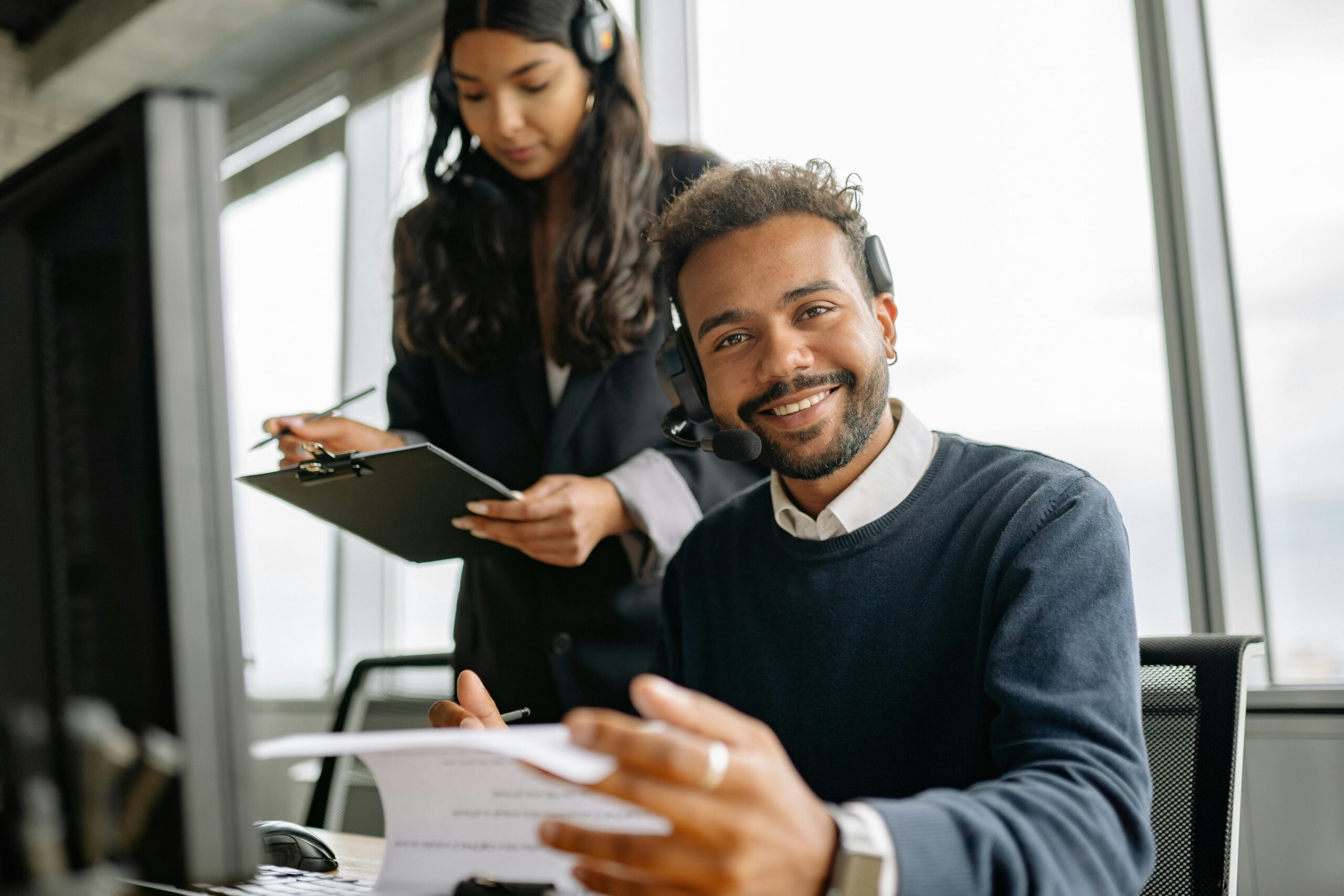 SERVICES Friendly call center agent wearing a headset, talking and smiling at his desk with a colleague.